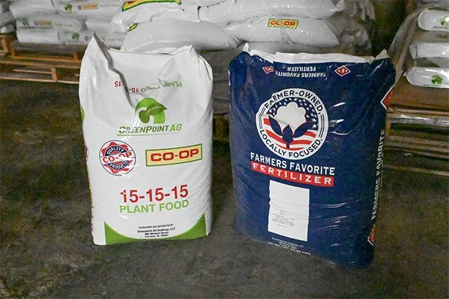Two large bags of fertilizer standing on a warehouse floor, one labeled 'CO-OP 15-15-15 Plant Food' and the other labeled 'Farmer-Owned Fertilizer'.