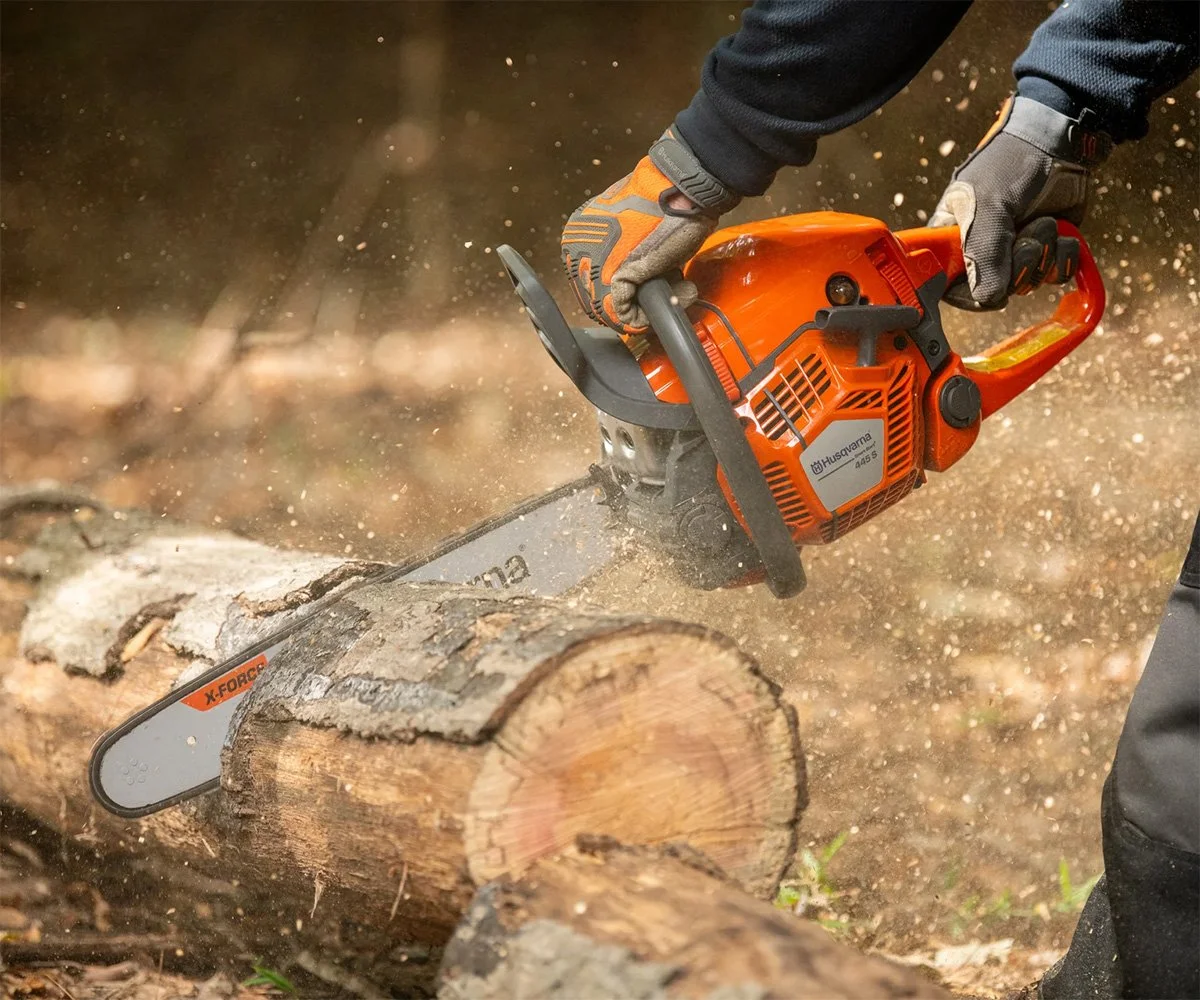 A person using an orange chainsaw to cut a log of wood outdoors, with sawdust flying around.