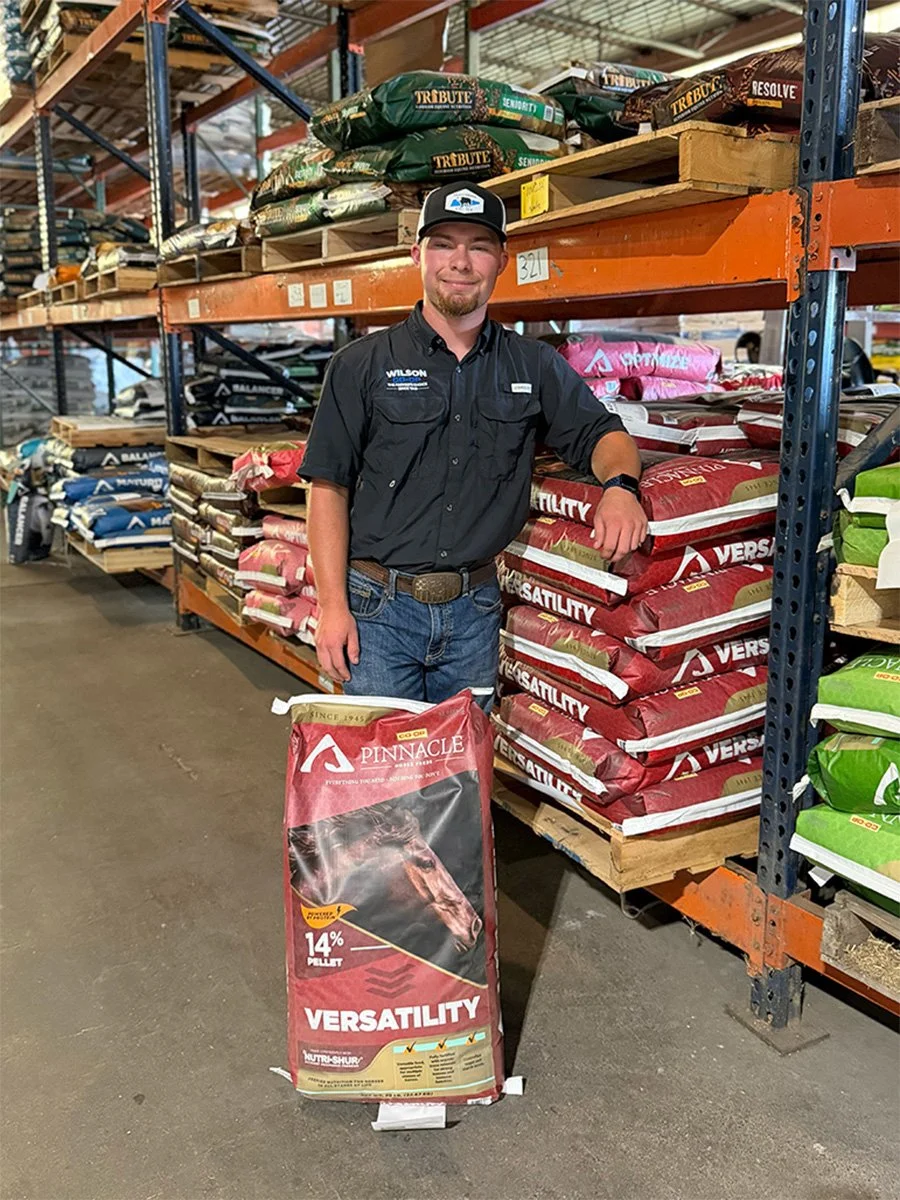 A man with a beard and mustache wearing a black button-up shirt and jeans stands in a warehouse aisle, holding a large bag of Pinnacle Versatility horse feed. Behind him are shelves stocked with bags of pet and animal feed in various colors and brands.