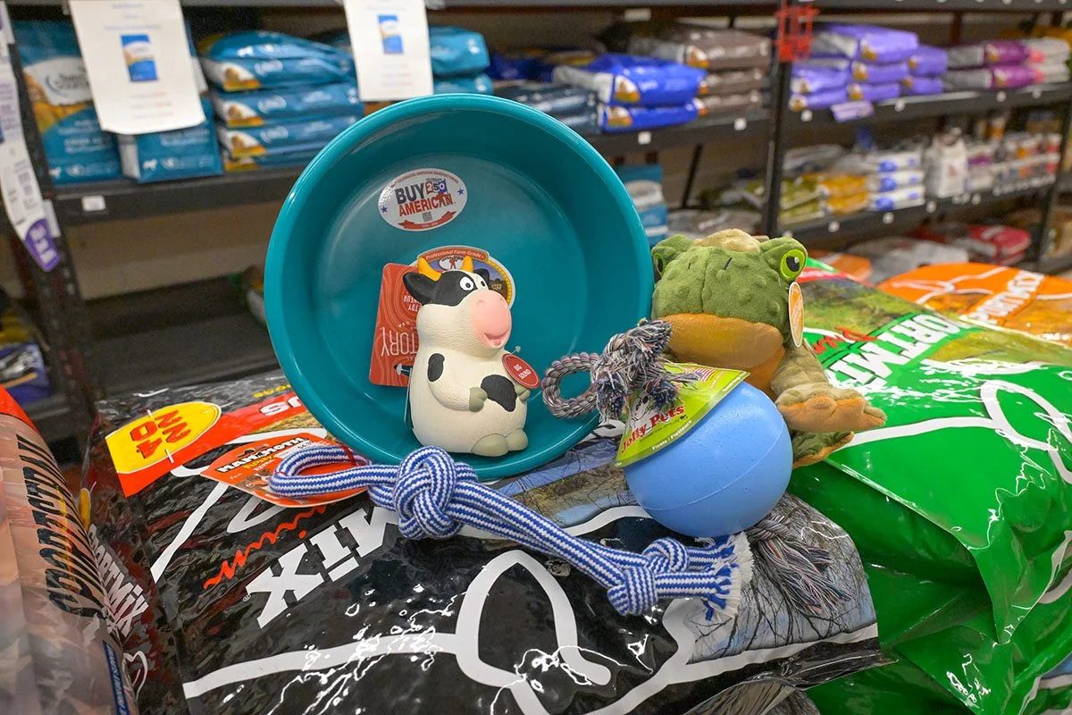 Pet supplies on store shelves, including a rubber cow toy, a frog plush toy, a blue ball, and a blue frisbee on top of large bags of pet food.