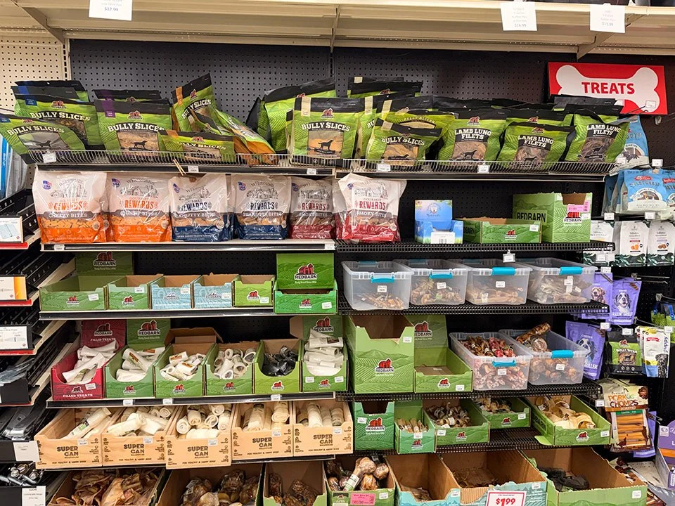 Dog treats and chew toys displayed on supermarket shelves, with bags of treats on the top and boxes of chews below.