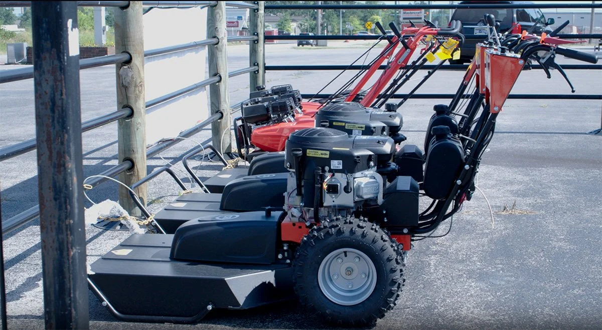 Multiple walk-behind lawn mowers lined up outside near a metal fence.