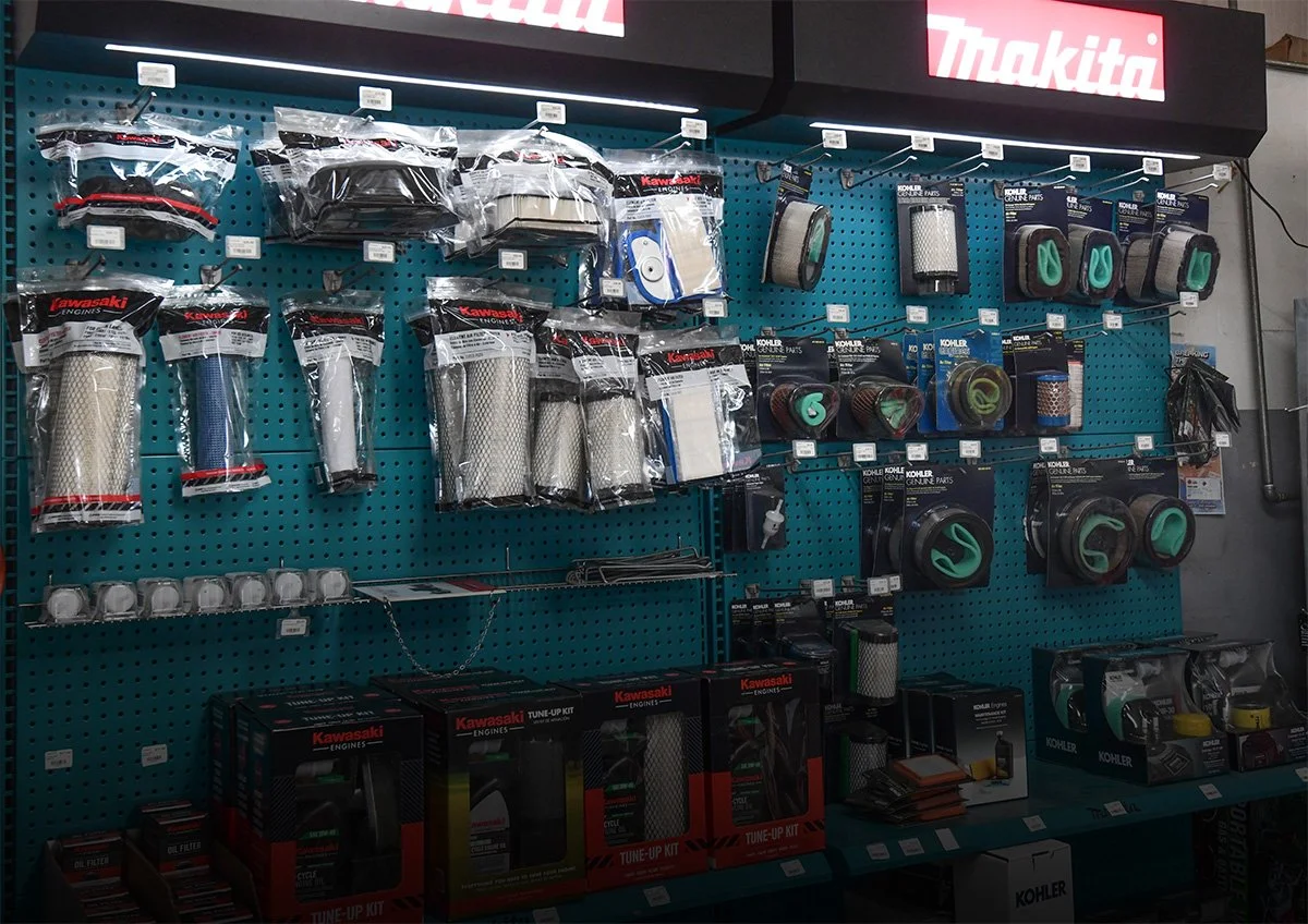 Automotive parts display on a teal pegboard wall, including filters, hoses, and radiator caps, with brand packaging from Kawasaki and Kohler, under a red and white Mazda sign.