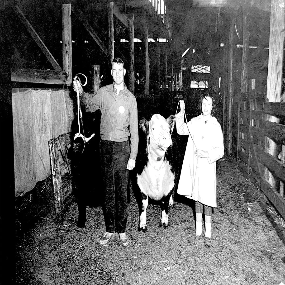Black and white photo of two women and a dog inside a barn. The women each hold a leash attached to the dog. The barn has wooden beams and shelves, with dirt ground.