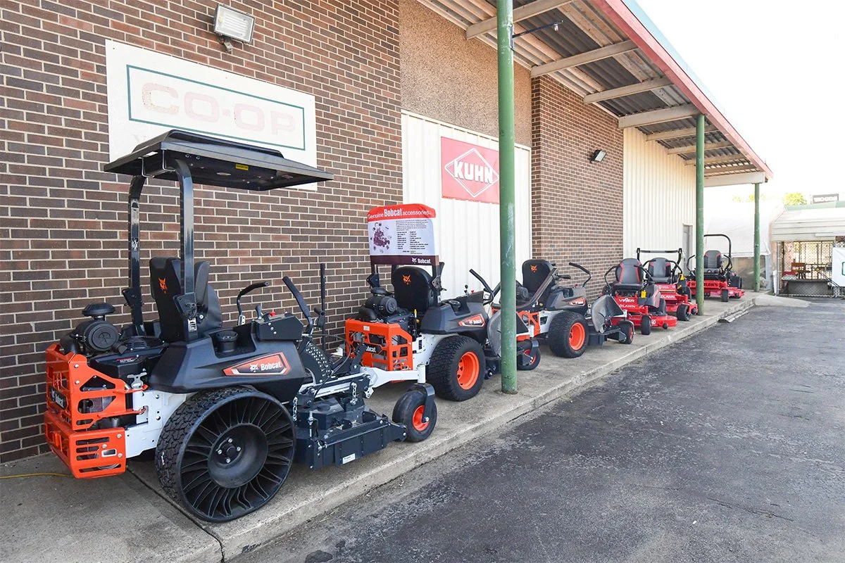 A row of various red and black Bobcat lawn and landscaping equipment lined up outside a brick building with a CO-OP sign and a KUHN sign.