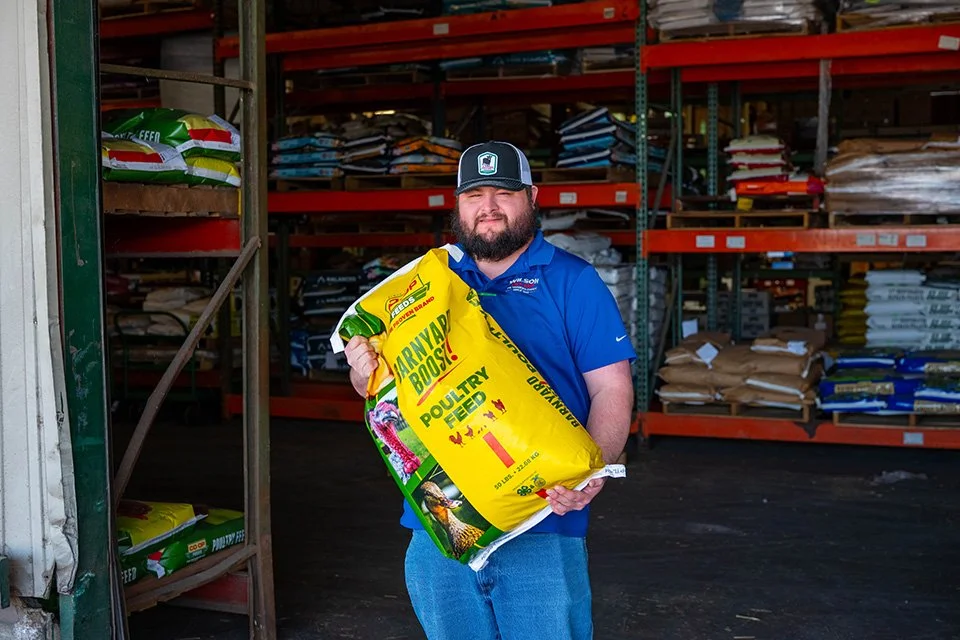 A man standing inside a store aisle holding a large yellow bag of chicken feed labeled 'Rooster Booster' with shelves of pet supplies in the background.