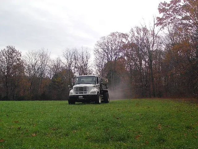 A white truck driving across a grassy field with dust behind it, surrounded by leafless trees on an overcast day.
