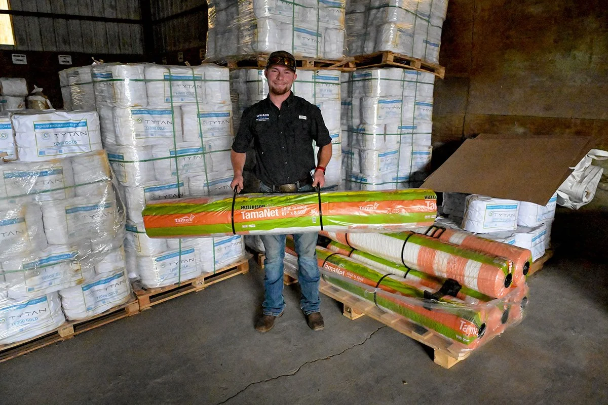 A man standing in a warehouse, smiling, holding a rolled up tarp labeled TamNet, with stacked packages of Tytan insulation around him.