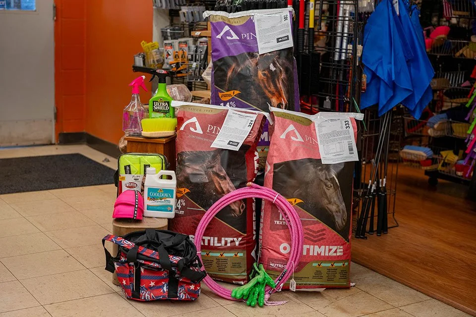 Display of pet supplies including bags of horse feed, grooming spray, coolant, a pink leash, gloves, and a first aid kit, with grooming tools and accessories on a wire rack in the background.
