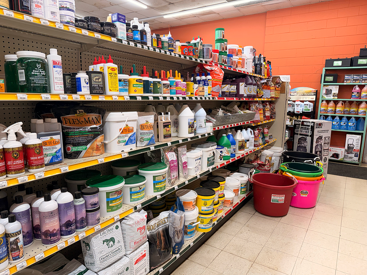 Aisle in a store with shelves stocked with pet supplies, including bottles, buckets, and bags of animal feed or supplements.