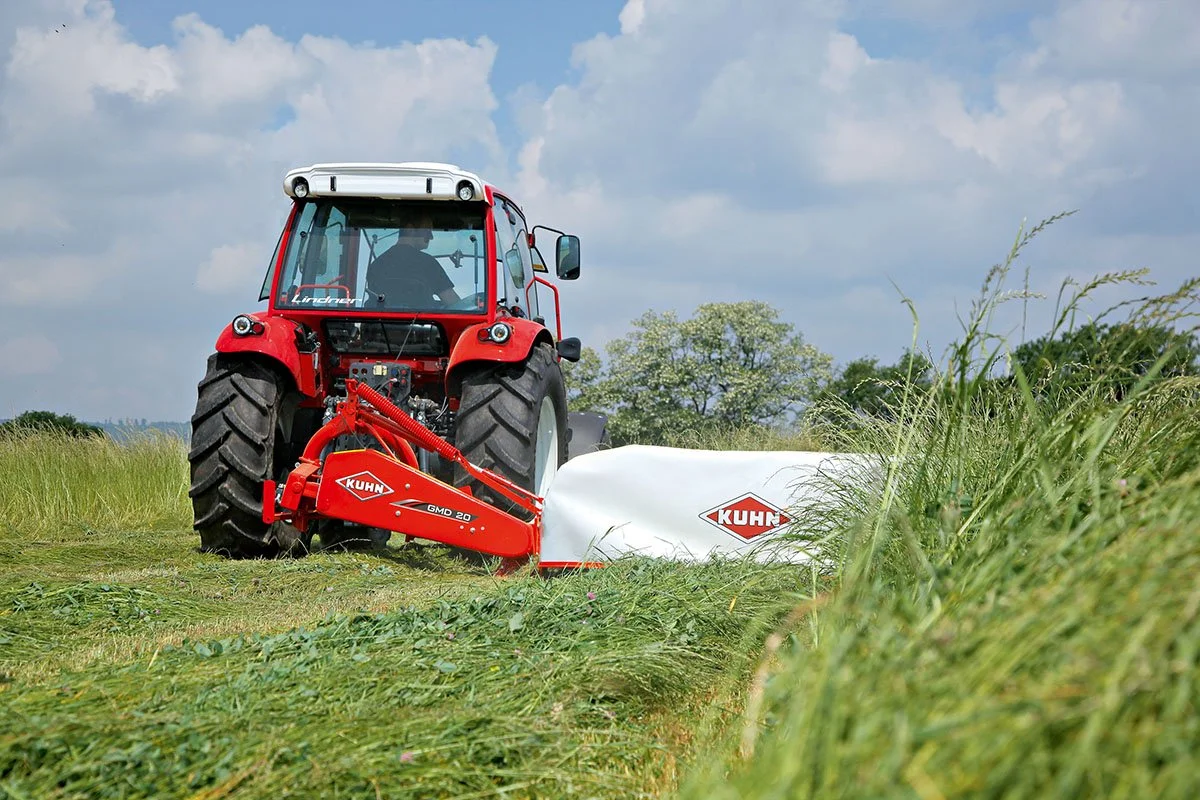 A red tractor with a KUHN abla mowing grass in a field with green trees and a cloudy sky in the background.