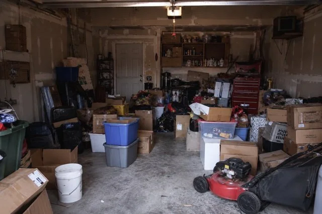 Garage filled with moving boxes, storage containers, and miscellaneous items, with a workbench and shelves in the background.