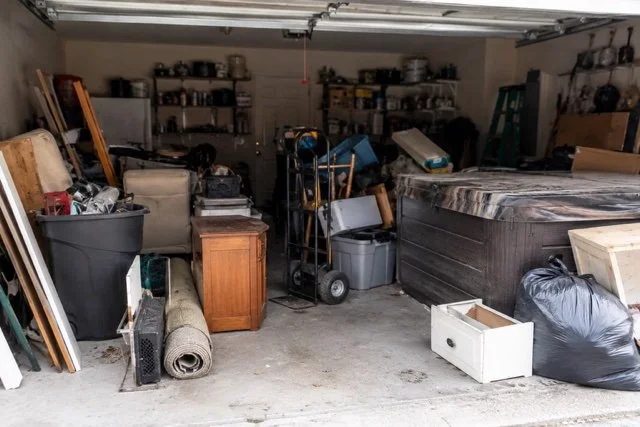 Garage filled with clutter including boxes, furniture, a mattress, storage bins, tools, and other household items.