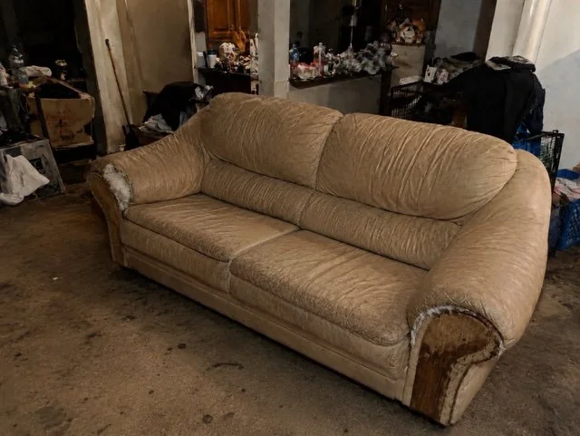 An old beige sofa with torn upholstery on the armrests, in a cluttered room with miscellaneous items around.