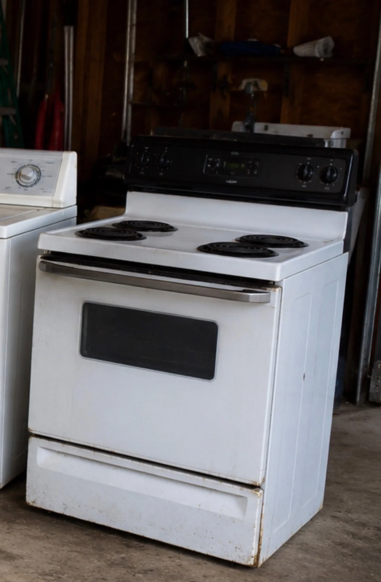 A white electric stove with four coil burners, a black control panel, and a self-clean oven, in a garage with wooden walls and concrete floor.