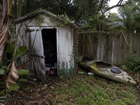 A weathered wooden shed with an open door surrounded by overgrown grass and trees, with an abandoned kayak lying on the ground nearby.