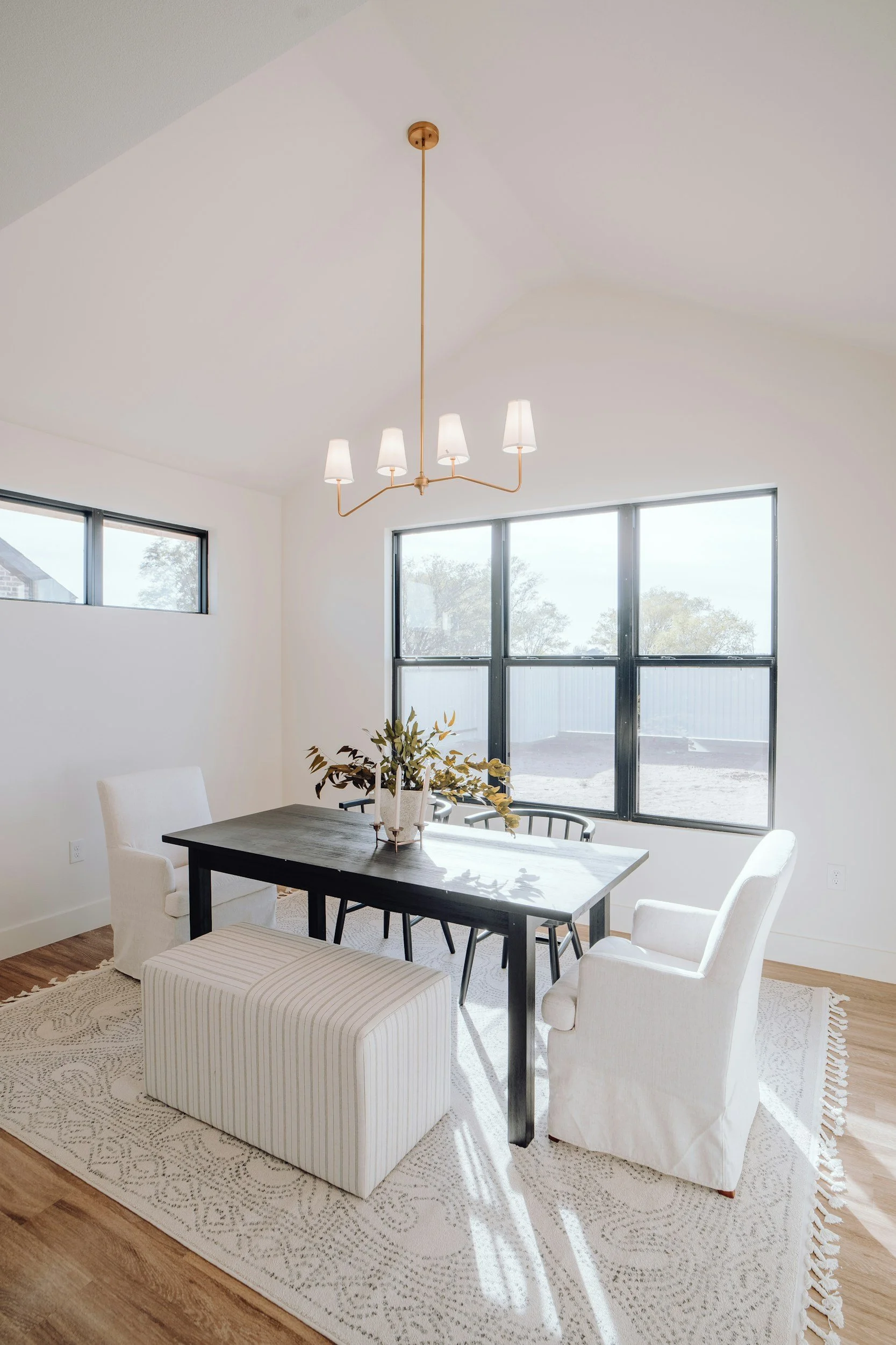 A bright dining room with large windows, a dark wooden table, white upholstered chairs, a beige patterned rug, a white cushioned bench, a centerpiece with plants, and a modern chandelier with five white lampshades hanging from a gold fixture.