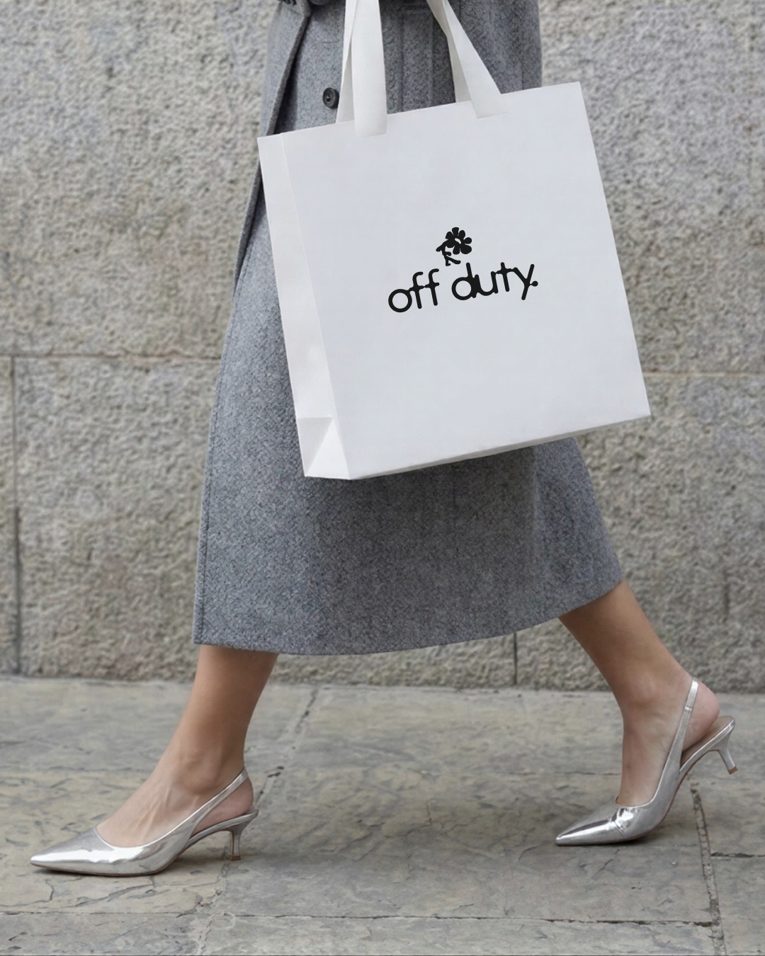 Une personne portant des chaussures à talons argentés, une jupe grise, une veste grise et tenant un sac blanc avec l'inscription "off duty" et un dessin de fleur.