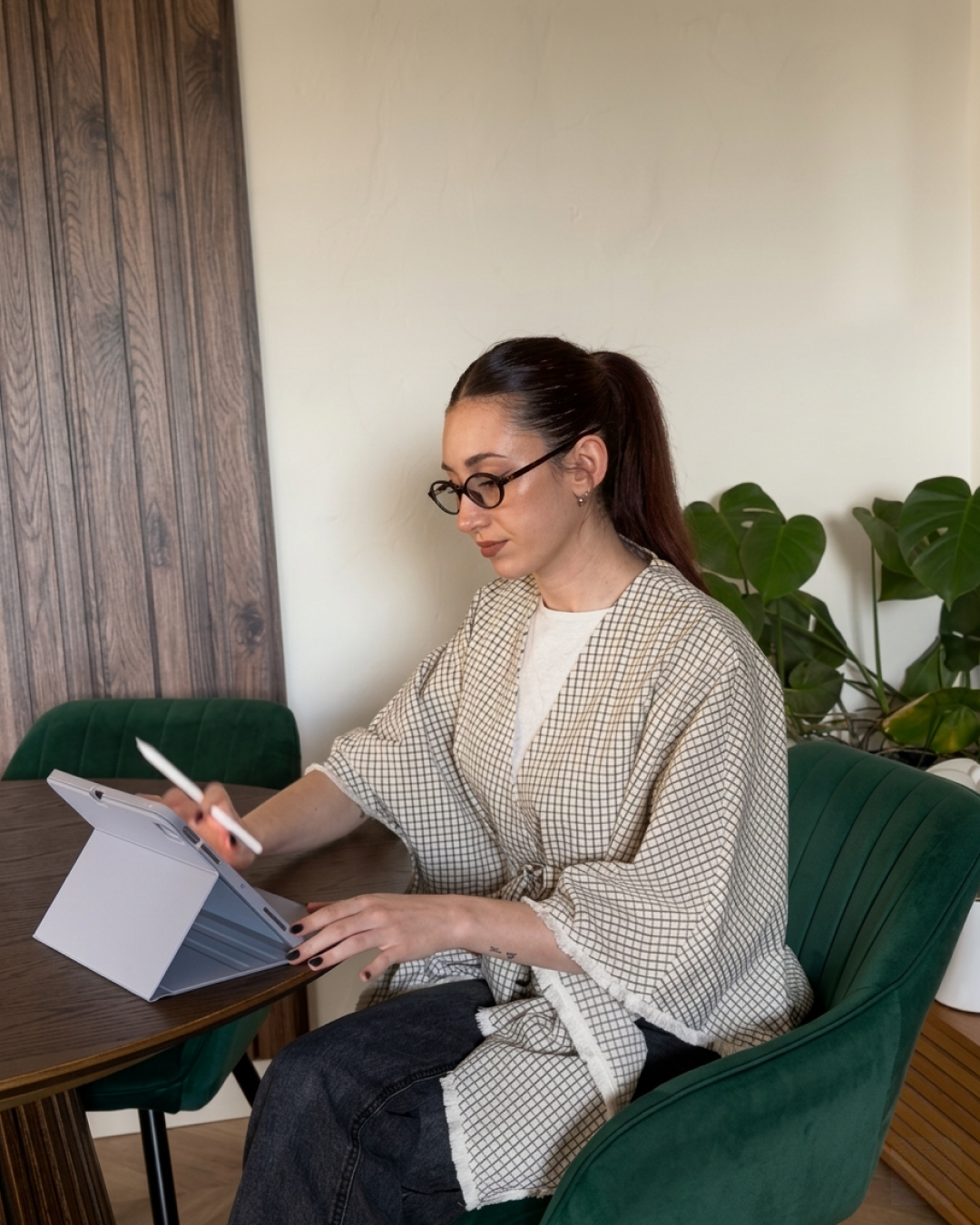 Femme assise à une table, utilisant une tablette numérique avec stylet, portant des lunettes, dans un environnement intérieur avec plantes vertes.