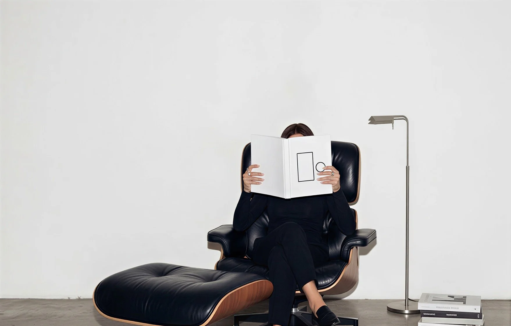 Une femme assise dans un fauteuil noir en cuir, lisant un livre blanc avec des dessins de formes géométriques, dans une pièce minimaliste avec un mur blanc, une lampe de lecture moderne à droite, et des magazines empilés au sol.