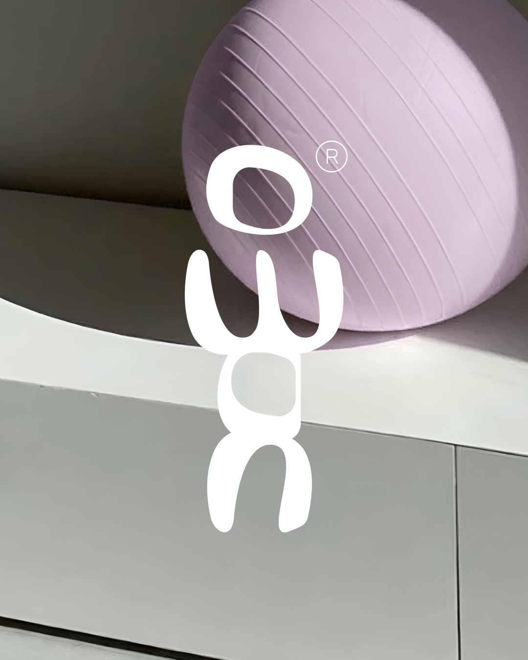 Gros plan sur un ballon de Pilates de couleur lilas orné du logotype 'noma' en lettres blanches arrondies et organiques. L'ensemble est posé sur une surface crème dans un intérieur épuré.