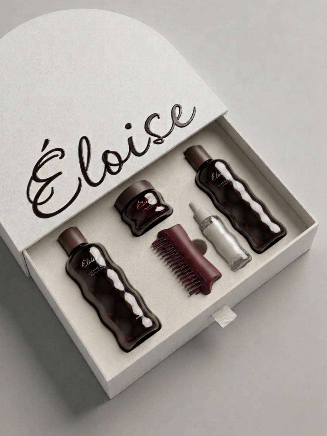 Photographie aérienne d'un coffret cadeau ouvert de couleur crème, posé sur une surface grise. L'intérieur du couvercle porte l'inscription "Éloise" en grande typographie manuscrite bordeaux. La base du coffret contient, dans un insert moulé, cinq pr