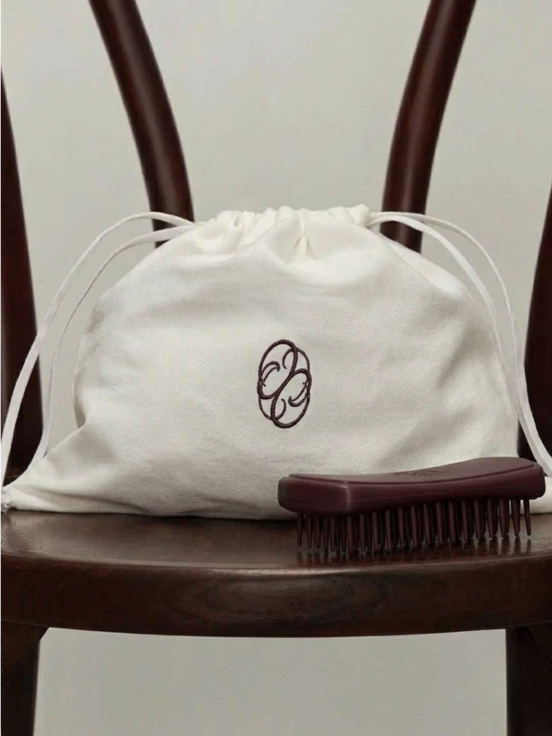 Photographie posée sur une chaise en bois foncé. Au centre, un pochon en tissu blanc cassé à cordon de serrage, brodé en son milieu du monogramme bordeaux de la marque Éloise. Juste devant, posée sur l'assise, une brosse de massage pour animal de for