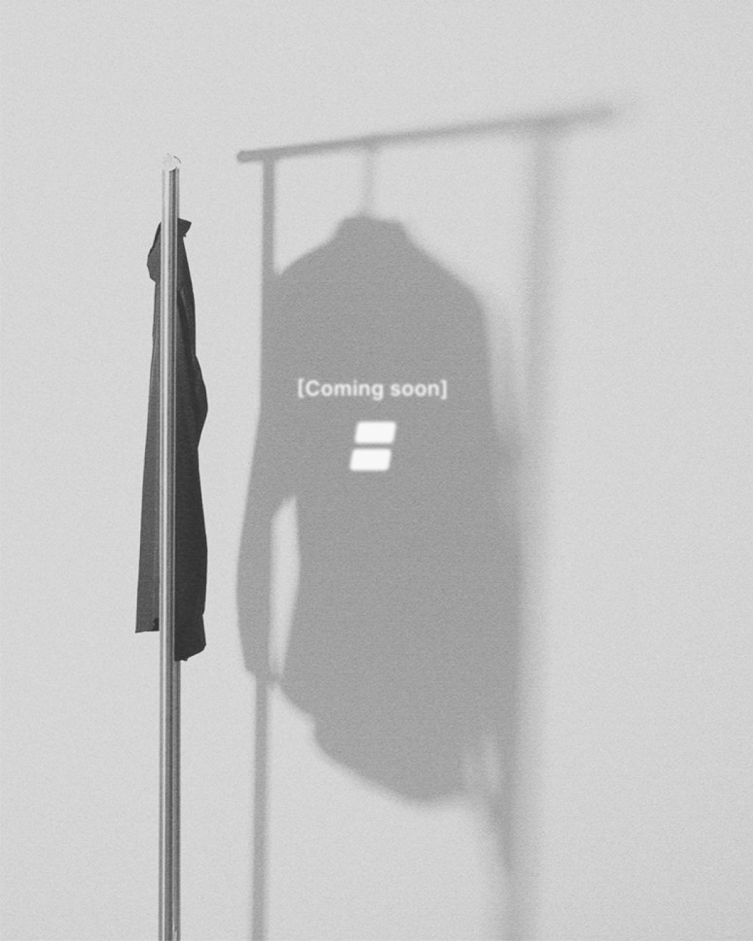 Une ombre portée d'un vêtement suspendu sur un mur blanc avec le texte "Coming soon" et un symbole, avec une veste suspendue à un portemanteau.