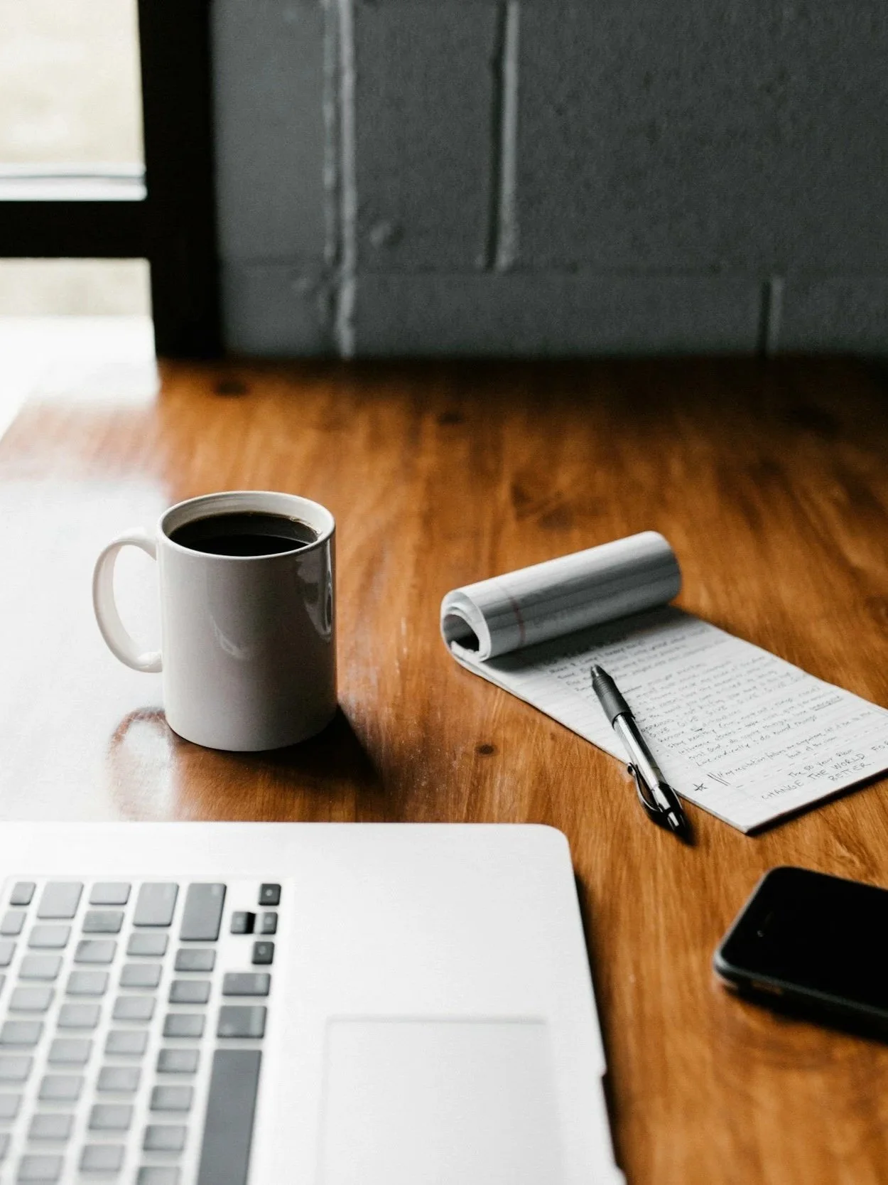 A desk with a white coffee mug filled with black coffee, a notepad with handwritten notes, a pen, a laptop keyboard, and a smartphone on a wooden surface, with a gray brick wall in the background.