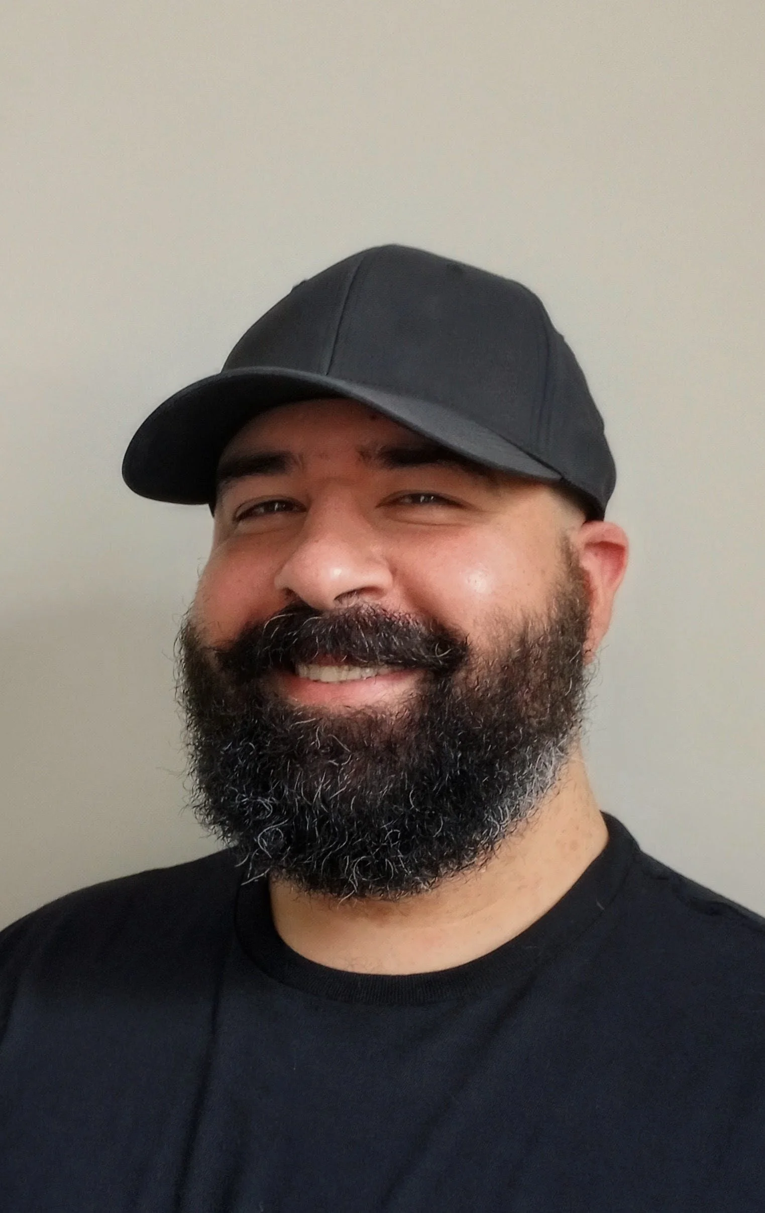 A man with a full beard and mustache, smiling, wearing a black baseball cap and black T-shirt, standing against a plain white wall.