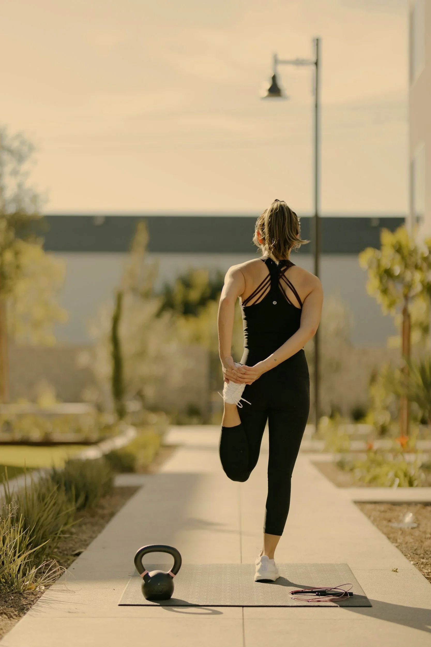 A woman stretching on a yoga mat outdoors during early morning or late afternoon, with kettlebell and jump rope nearby.