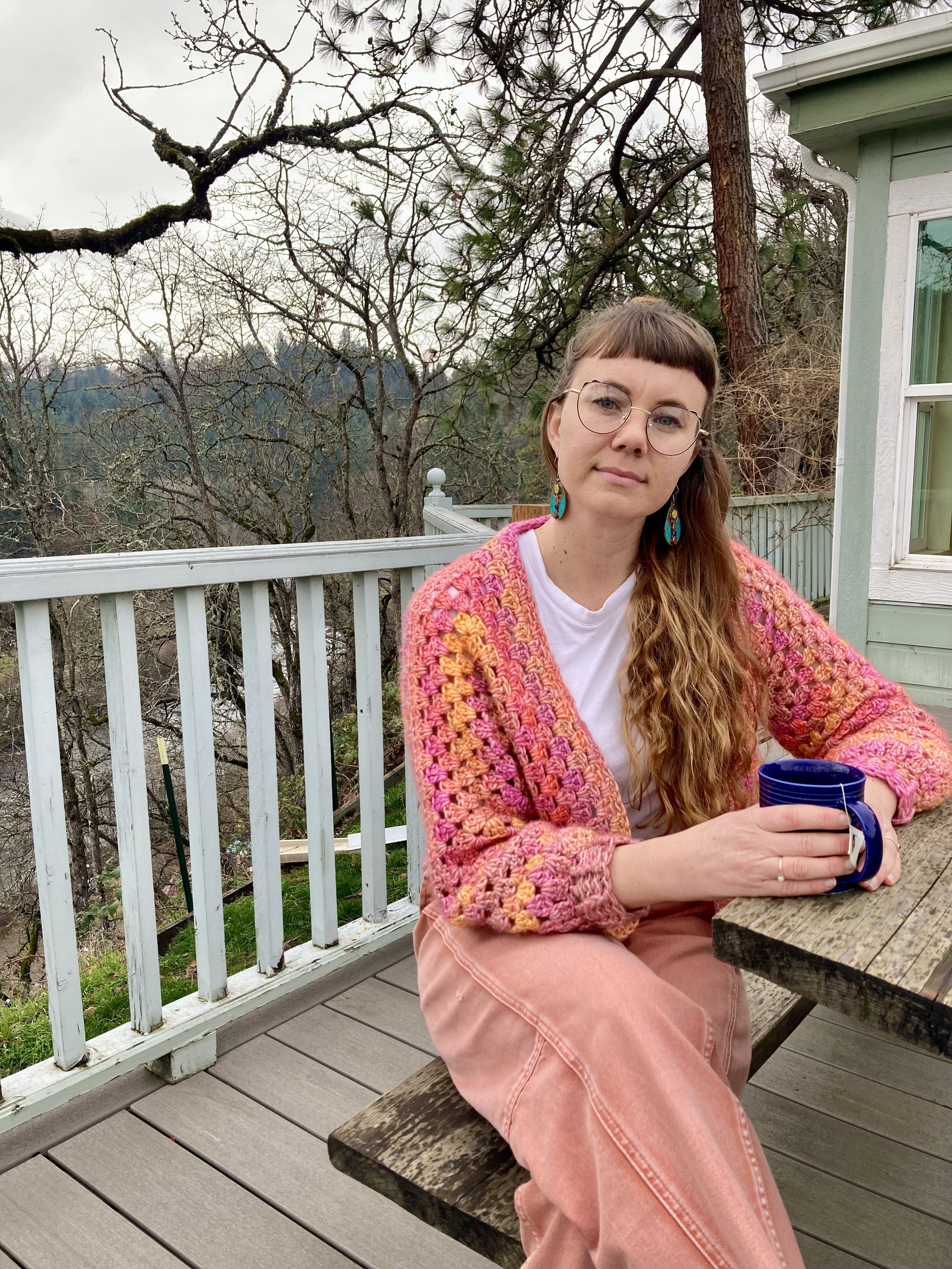 Woman sitting on a wooden bench on a porch holding a blue mug. She wears glasses, large colorful earrings, a pink crochet cardigan, and pink pants. There is a white railing, a house with a light green exterior, and leafless trees in the background.