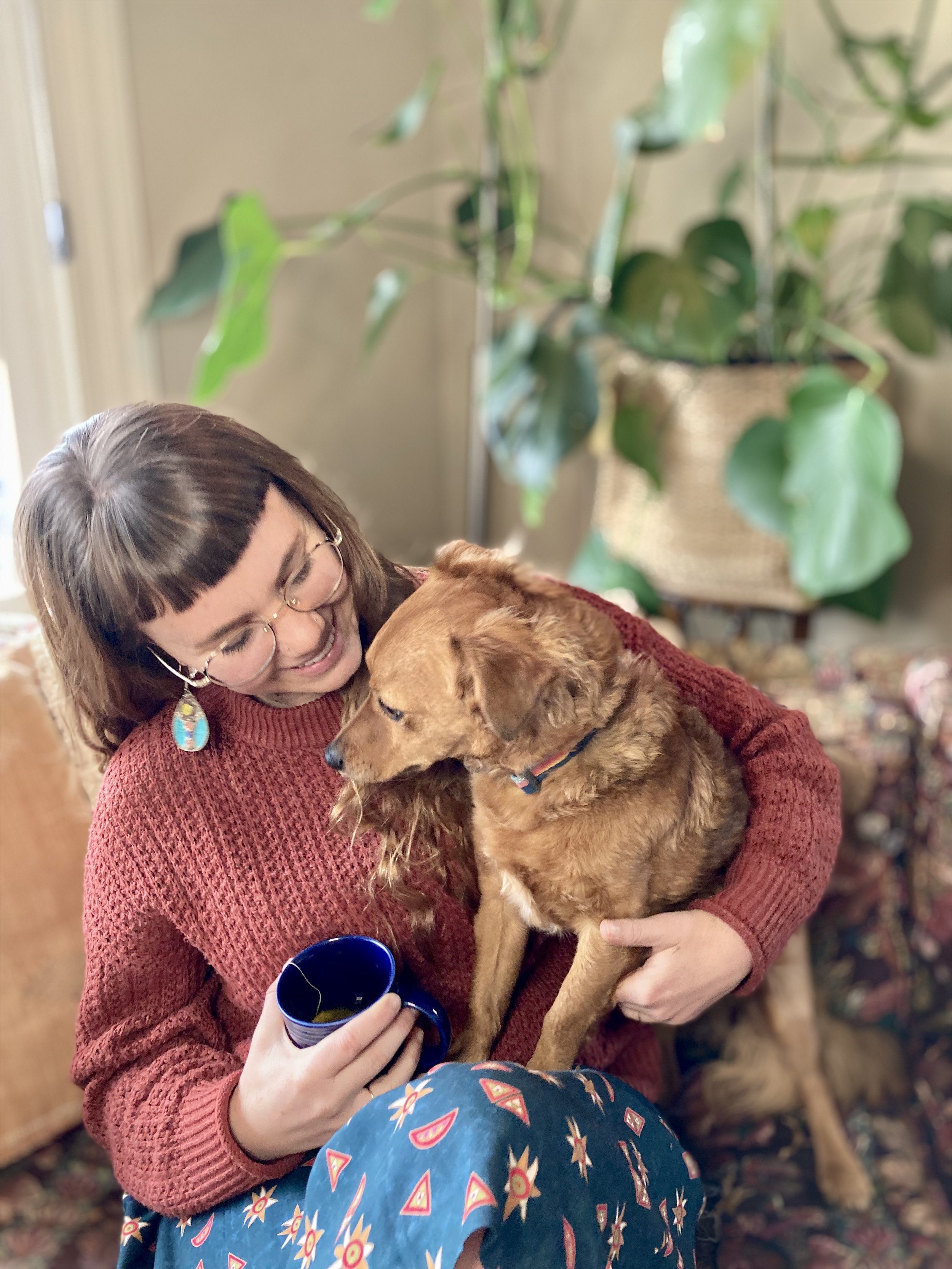 A woman with glasses and a red sweater smiling while holding a brown dog, with a large green potted plant in the background.