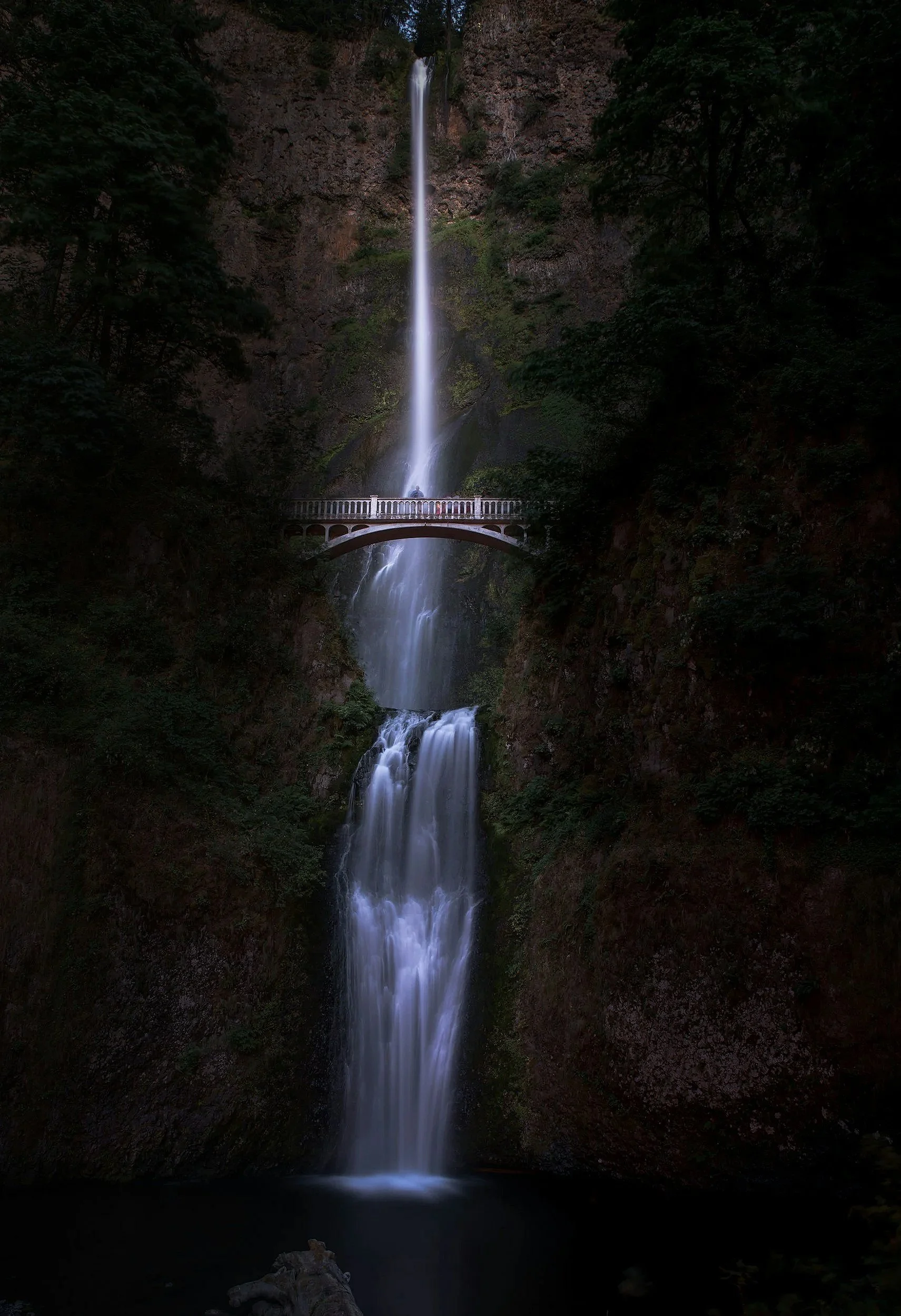 A multi-tiered waterfall flowing down a rocky cliff surrounded by green trees, with a small bridge crossing in front of the waterfall.