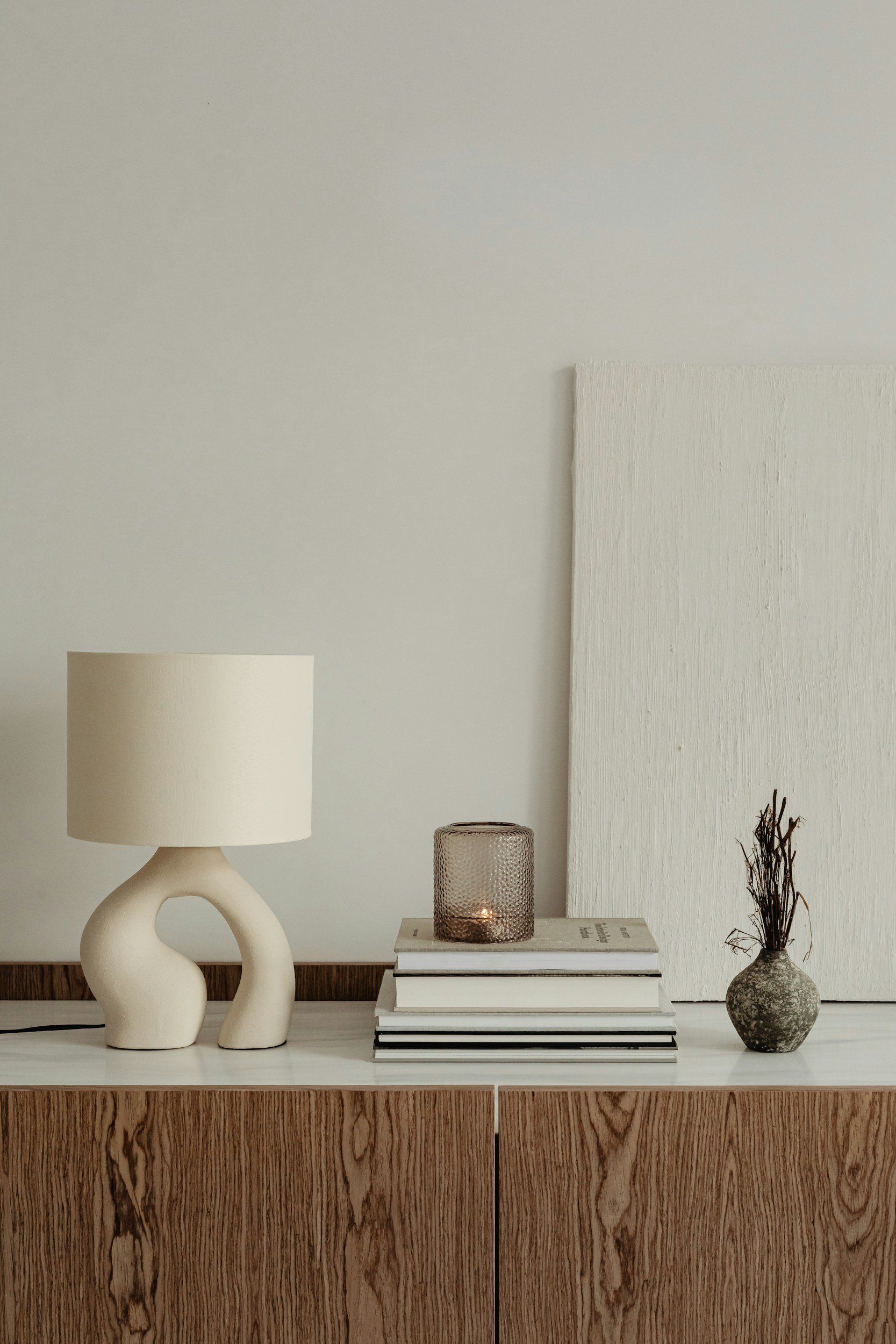 Simple interior decor setup featuring a white table with a ceramic lamp, a stack of books, a candle holder, and a vase with dried branches, all against a white wall with minimalistic artwork and decor.