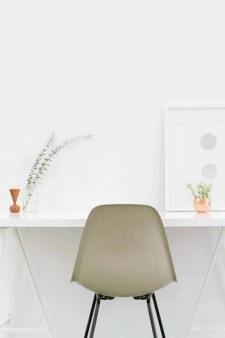 Minimalist white desk with beige chair, decorated with potted plants and framed minimalist art, in a bright modern room.