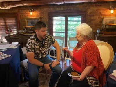 RJ Shaw of Galveston Unscripted speaks with DRT member at Sidney Sherman DRT meeting at the Bryan Museum in a cozy room with brick walls, wooden window blinds, and warm lighting.