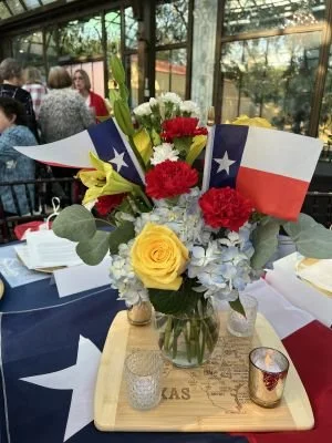 A floral arrangement with yellow roses, red carnations, blue hydrangeas, and greenery, featuring Texas and patriotic flags, placed on a wooden tray with candles, set on a table at a gathering with people in the background.