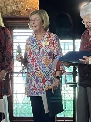 A new Sidney Sherman Chapter member holding a bluebonnet in one hand and her certificate in the other, standing indoors with sunlight coming through the window behind her, and others nearby.