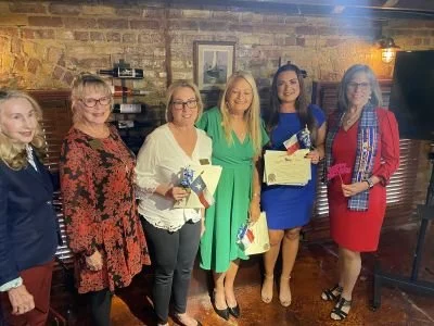 Six women standing together indoors, holding gifts, in a restaurant or cafe setting with exposed brick walls and warm lighting.