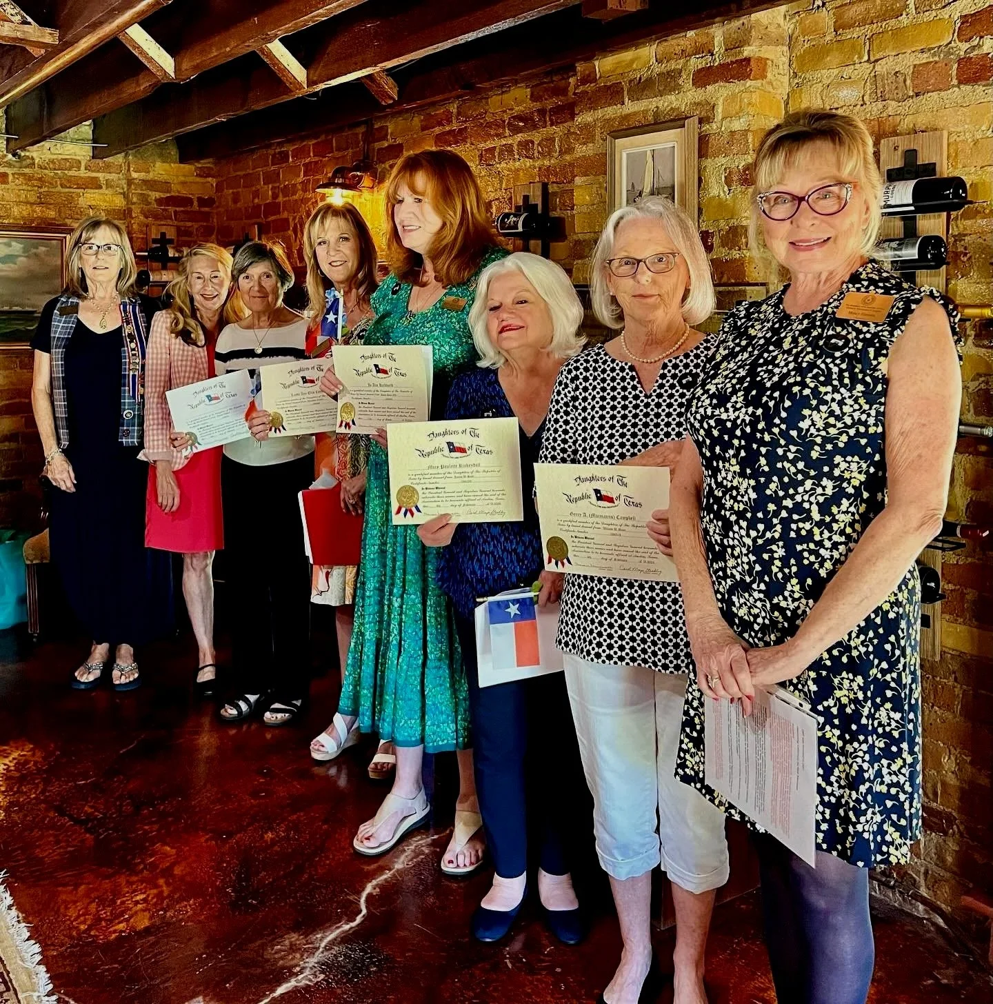 New Sidney Sherman Chapter DRT members induction.  Women standing indoors, holding certificates and small Texas flags, in front of a brick wall and framed artwork.