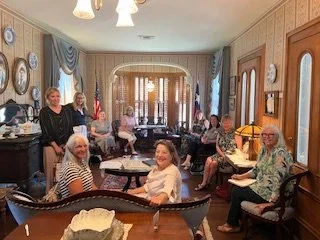 The Cradle Committee.  Group of women gathered at the Cradle, a cozy, Victorian-era room with floral wallpaper, antique furniture, and decorative plates on the wall.  The birthplace of the DRT