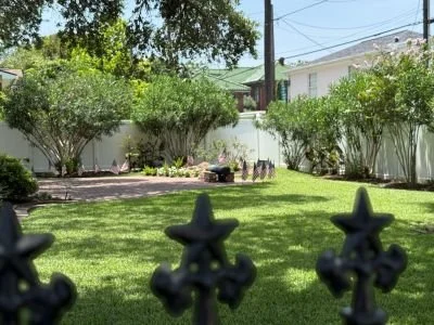 View of the Cradle backyard with a lush green lawn, surrounded by bushes and trees, with decorative black metal fence in the foreground, and a blue sky in the background.