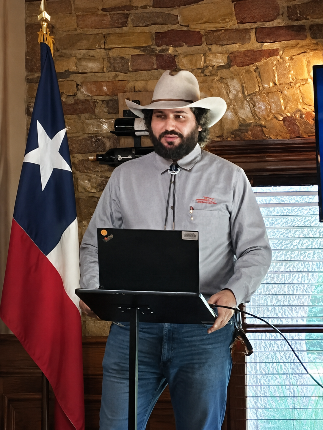 A man dressed as a cowboy, wearing a large cowboy hat, a collared shirt, and jeans, standing behind a lectern with a laptop, in front of the Texas state flag and a stone wall interior.