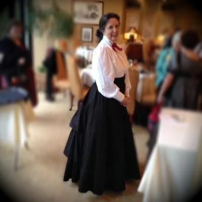 A woman dressed in a historical or period costume standing in a room with several other people in the background.