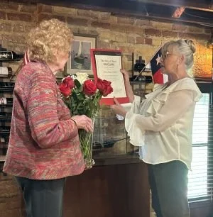 Two women exchanging flowers and a framed certificate at a Sidney Sherman DRT meeting