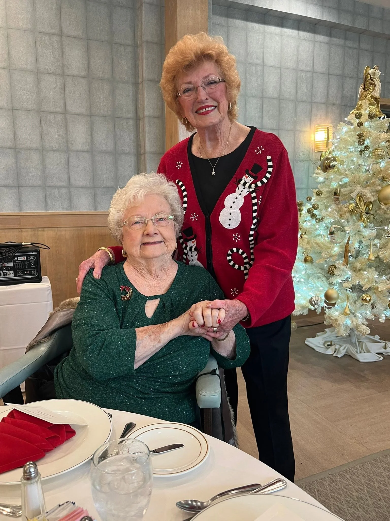DRT members at the Christmas Party An elderly woman sitting and smiling, holding hands with a middle-aged woman standing next to her, both dressed festively with Christmas decorations and a white Christmas tree in the background.