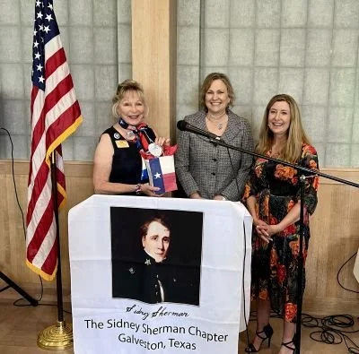 DRT members and State Rep Teri Leo Wilson standing behind a podium, the American flag in the background, and a banner that reads 'The Sidney Sherman Chapter Galveston, Texas'.