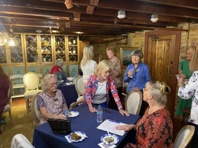 A group of women gathered around a table at a DRT meeting at the Bryan Museum in Galveston in a rustic wooden room with brick walls. Some women are sitting, while others are standing, engaged in conversation.
