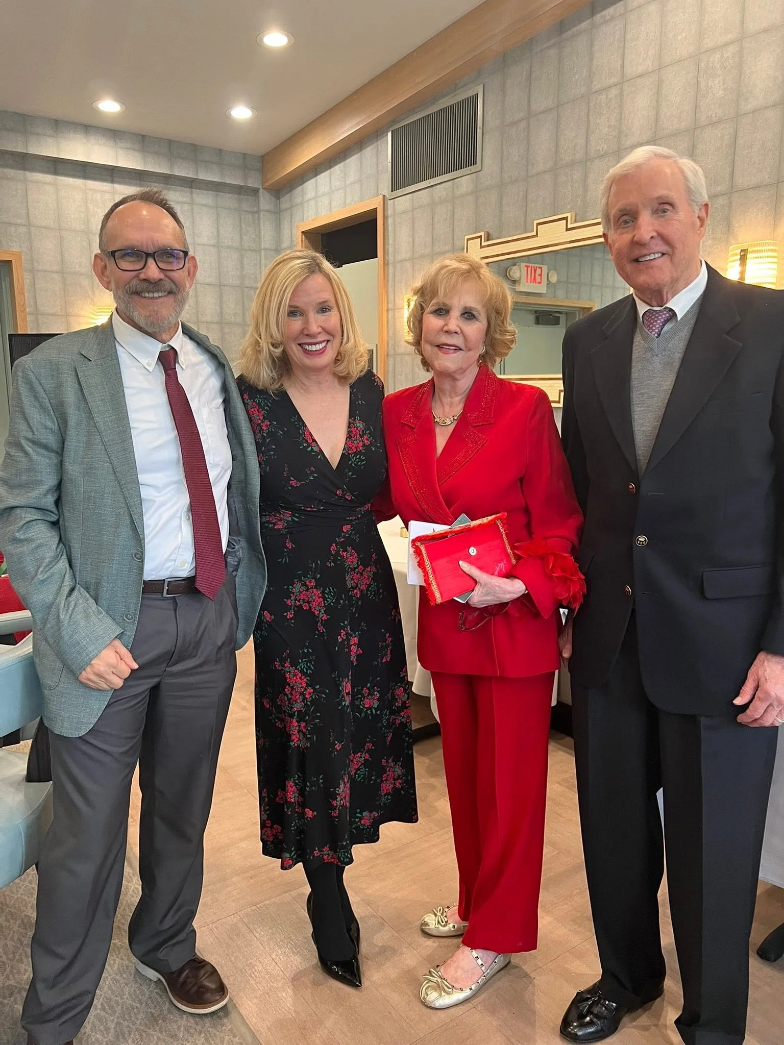 Four people standing together in a room, smiling at the camera. The group consists of two men and two women, with the woman in the red outfit holding a folder or book. The background includes a textured wall, a mirror, and an exit sign.